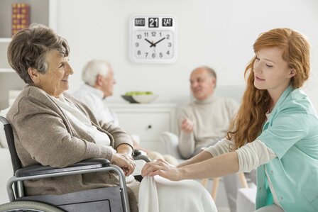 Dementieklok Fysic FK950 aan de muur in een zorginstelling met oudere vrouw in rolstoel en verzorgende