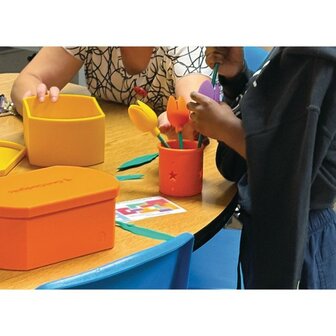 Twee mensen zitten aan een tafel en gebruiken de Geri-Gadgets Flower Bucket om kleurrijke bloemen in een oranje pot te plaatsen