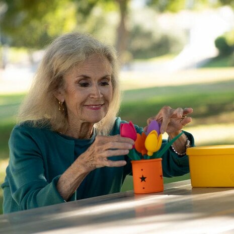Oudere vrouw plaatst kleurrijke siliconen bloemen in een oranje pot van de Geri-Gadgets Flower Bucket. Ontspannende activiteit 